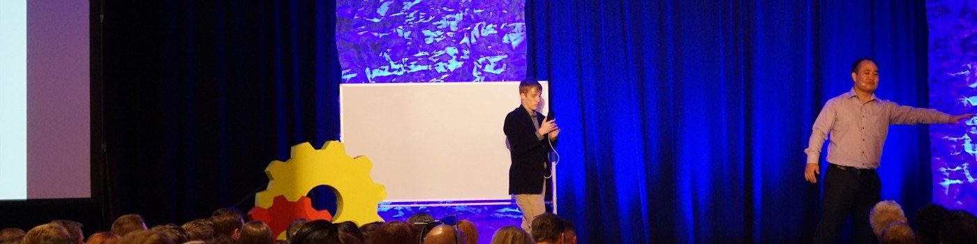 Book Dennis Yu to Speak — Keynotes & AI Agent Workshops 5 Dennis Yu presenting on a blue-lit conference stage, gesturing toward a co-presenter with the audience in front