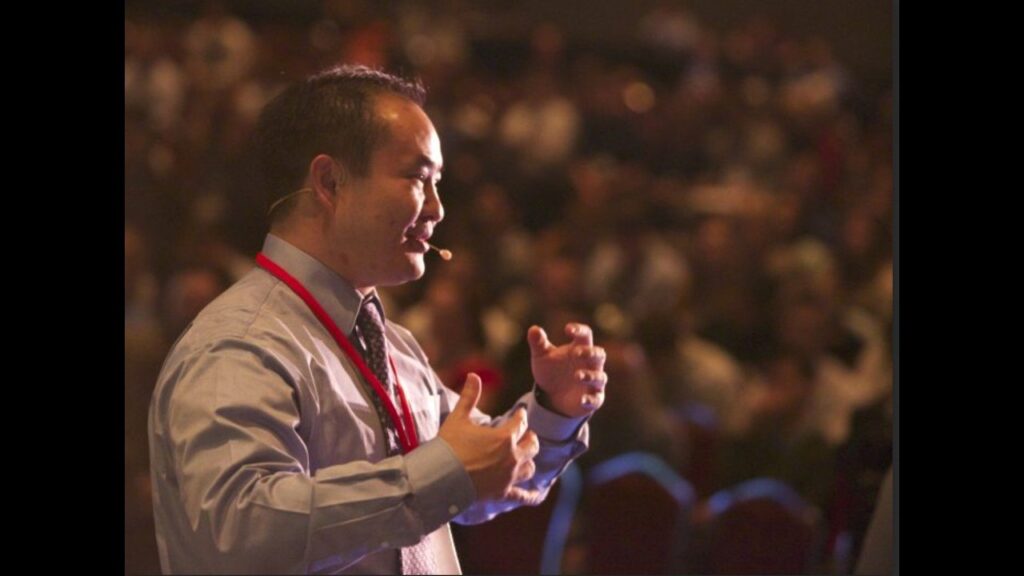 Dennis Yu speaking on stage with a headset microphone, gesturing, with a large audience filling the room behind him